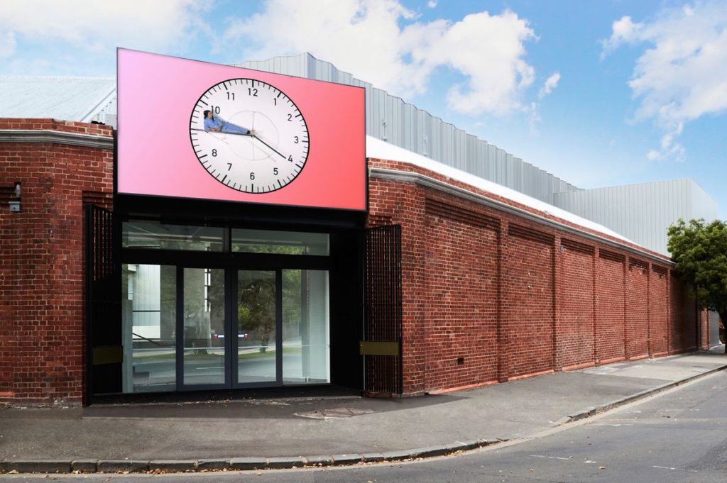 Image of Buxton Contemporary, a one-storey brick building. The building has a red built board/artwork showing a clock with a human lying on the big hand.