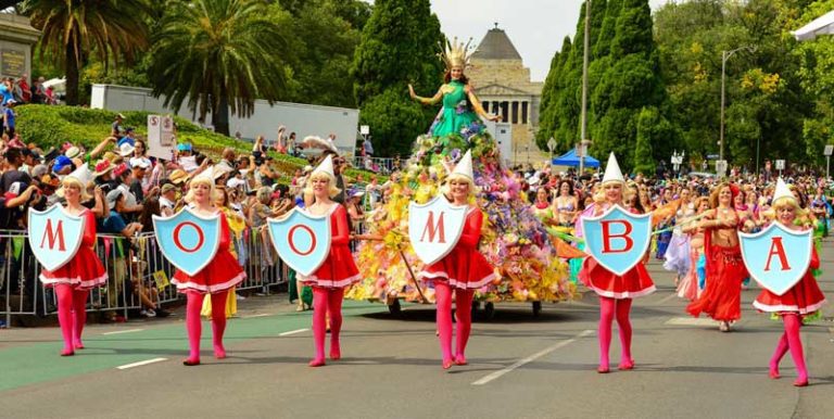 Image of Moomba parade. The focus is on 6 women dressed in red, all holding one letter each to spell out Moomba