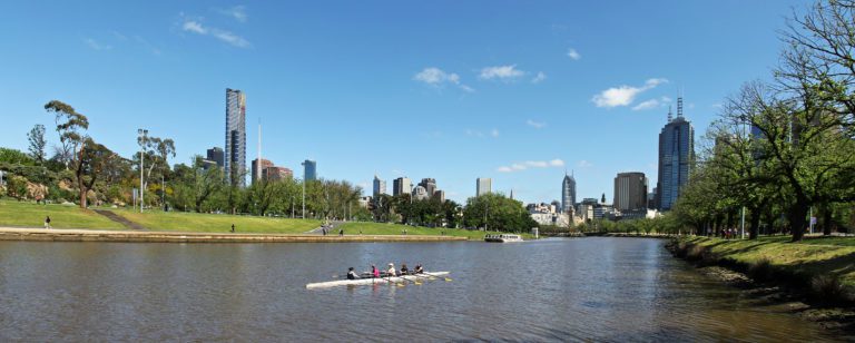 Image of the Yarra River with clear blue sky. The river has one rowboat on it and city buildings standing in the background.