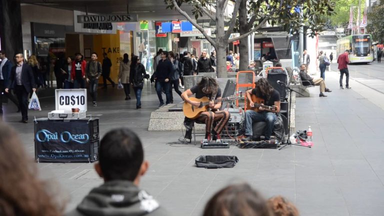 Image of two man on the Melbourne streets busking. Both men have long brown hair and are playing guitars.