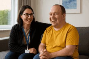 A smiling support worker and NDIS participant sitting together in a bright, modern living room, representing the trust and care of a registered SIL provider.